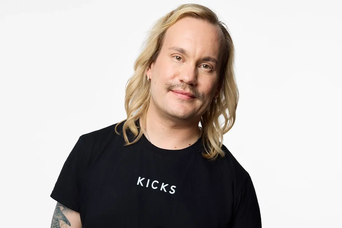 Henkilöllä on olkapäille ulottuvat vaaleat hiukset ja musta T-paita, jossa lukee valkoisella KICKS, vaaleaa taustaa vasten.
