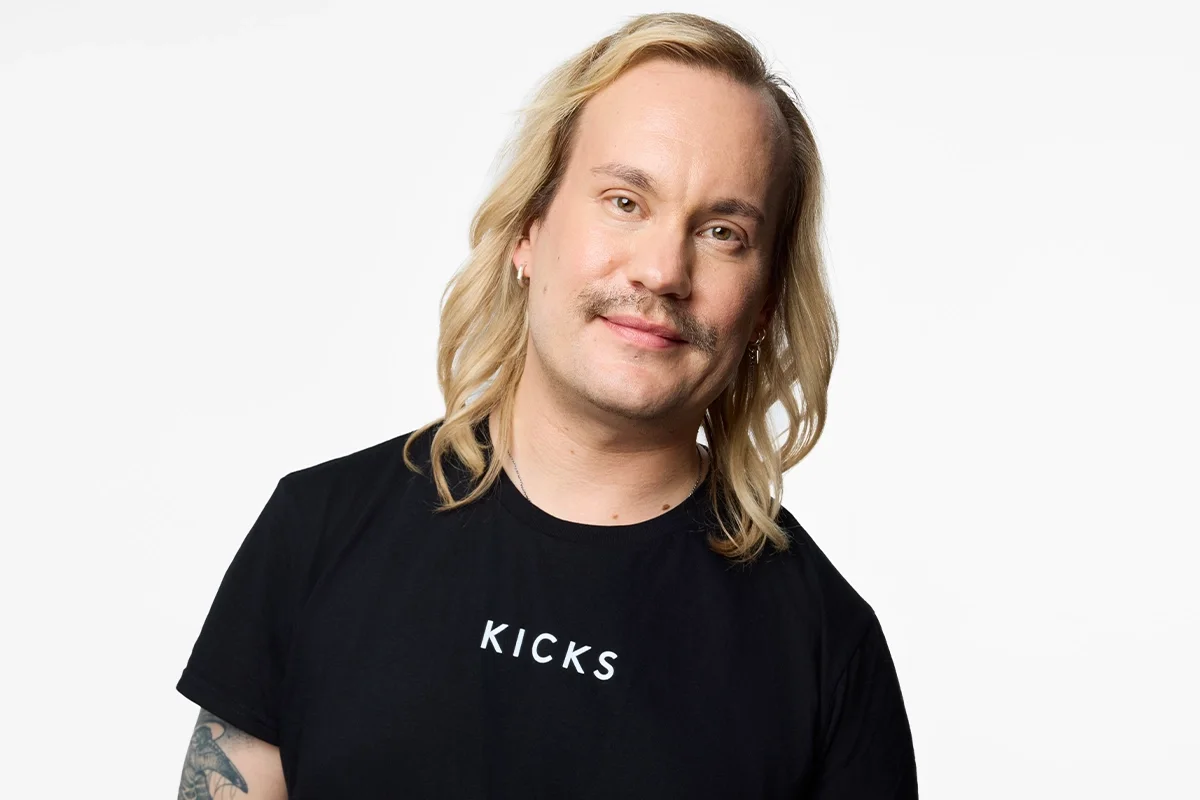 Henkilöllä on olkapäille ulottuvat vaaleat hiukset ja musta T-paita, jossa lukee valkoisella KICKS, vaaleaa taustaa vasten.