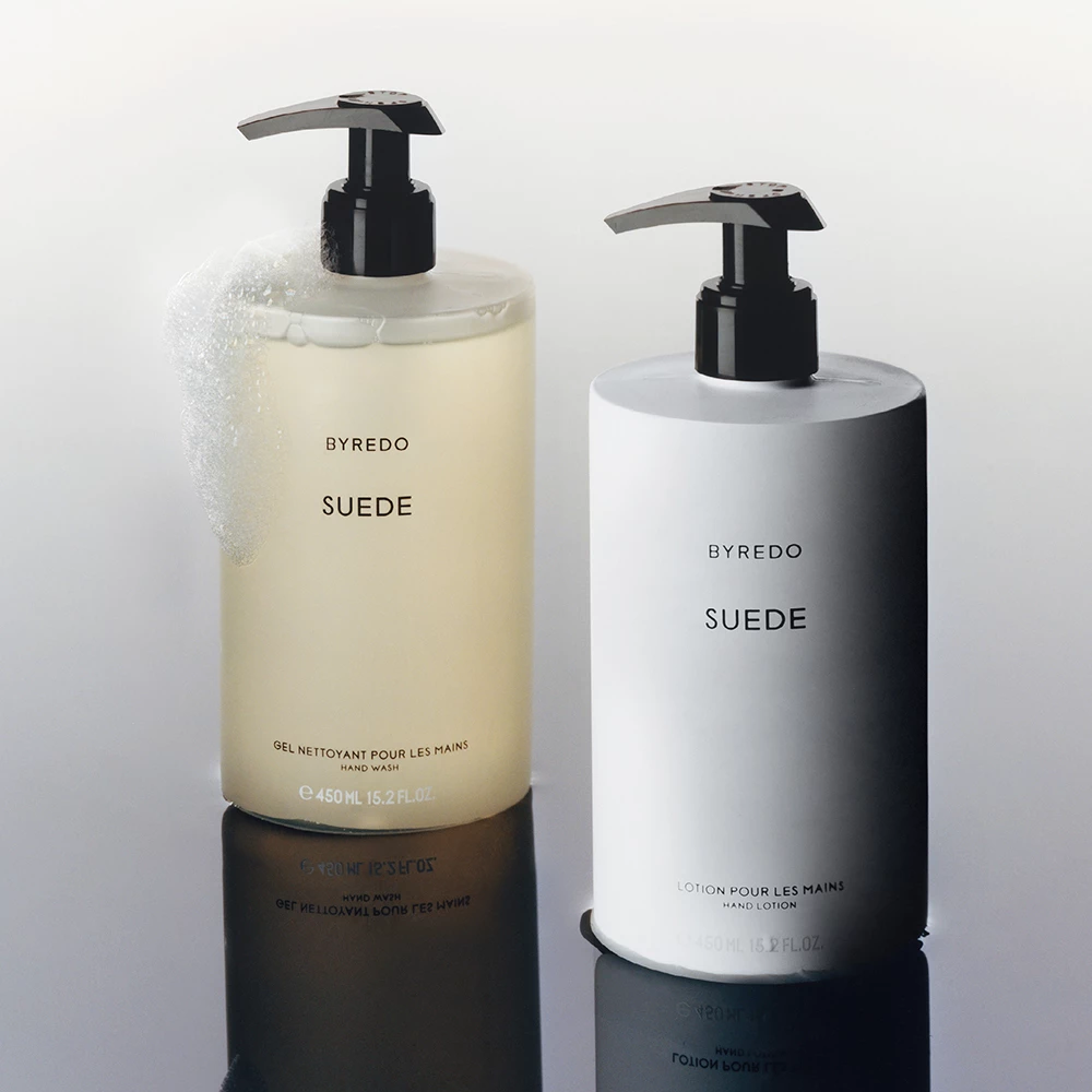 Byredo-produkter med namnet "Suede". Den ena är en handtvål i en genomskinlig flaska med en svart pump och skum på toppen, och den andra är en handkräm i en vit flaska med en svart pump. Bakgrunden är ljus och reflekterar produkterna.