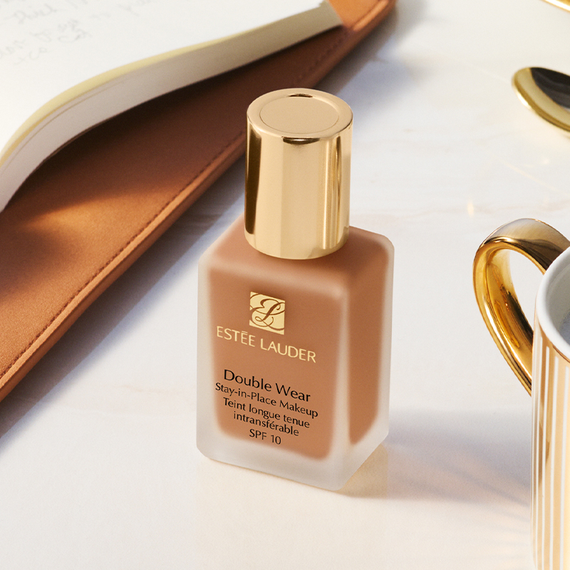 Estée Lauder Double Wear Stay-in-Place Makeup SPF 10 -meikkipullo marmoripinnalla kultareunaisen kupin ja avoimen muistikirjan vieressä.