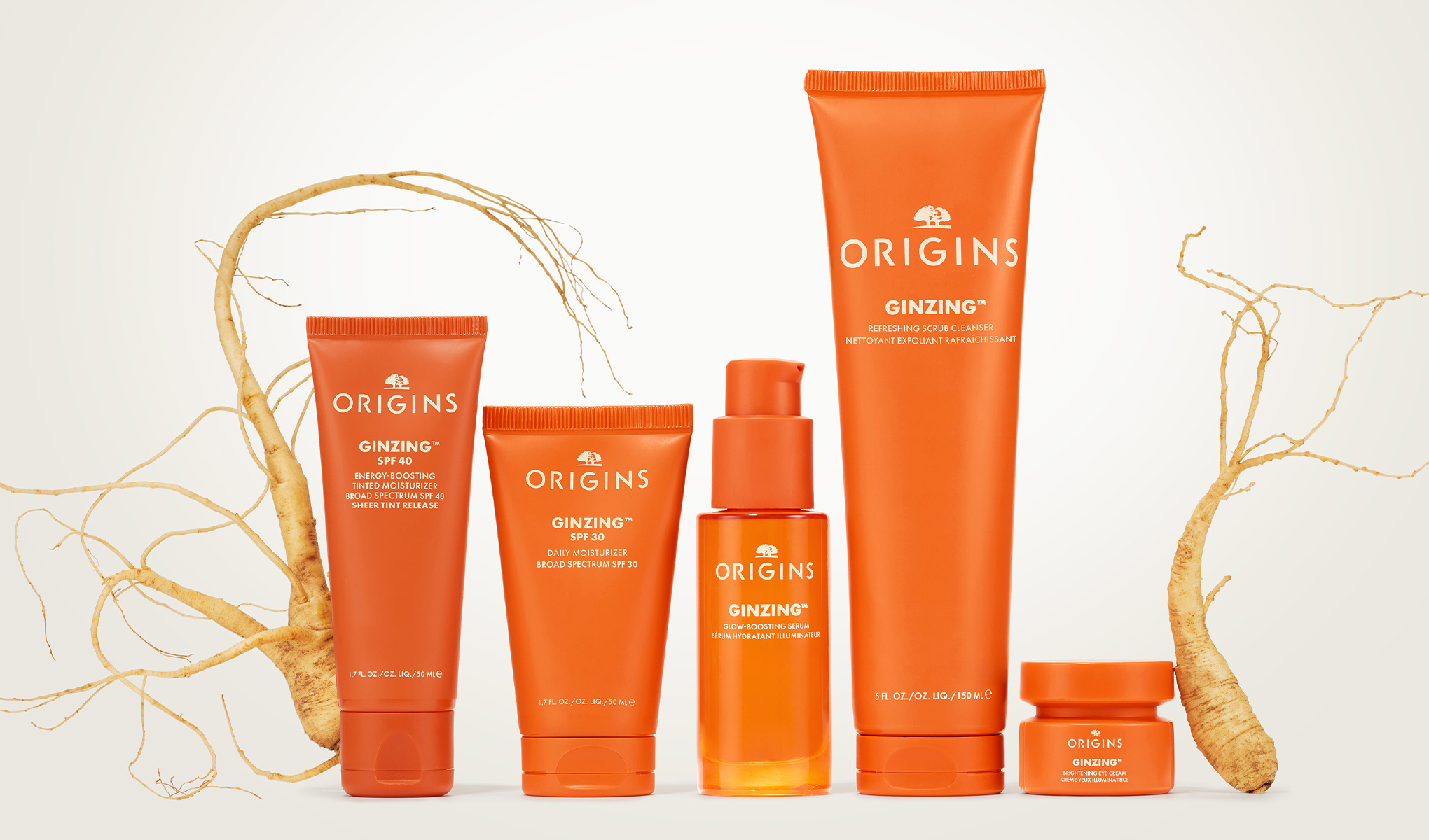 Origins GinZing -tuotteita, mukaan lukien kosteusvoiteet, seerumi ja kuorintavoide, aseteltuna valkoiselle taustalle ginseng-juurten kanssa.