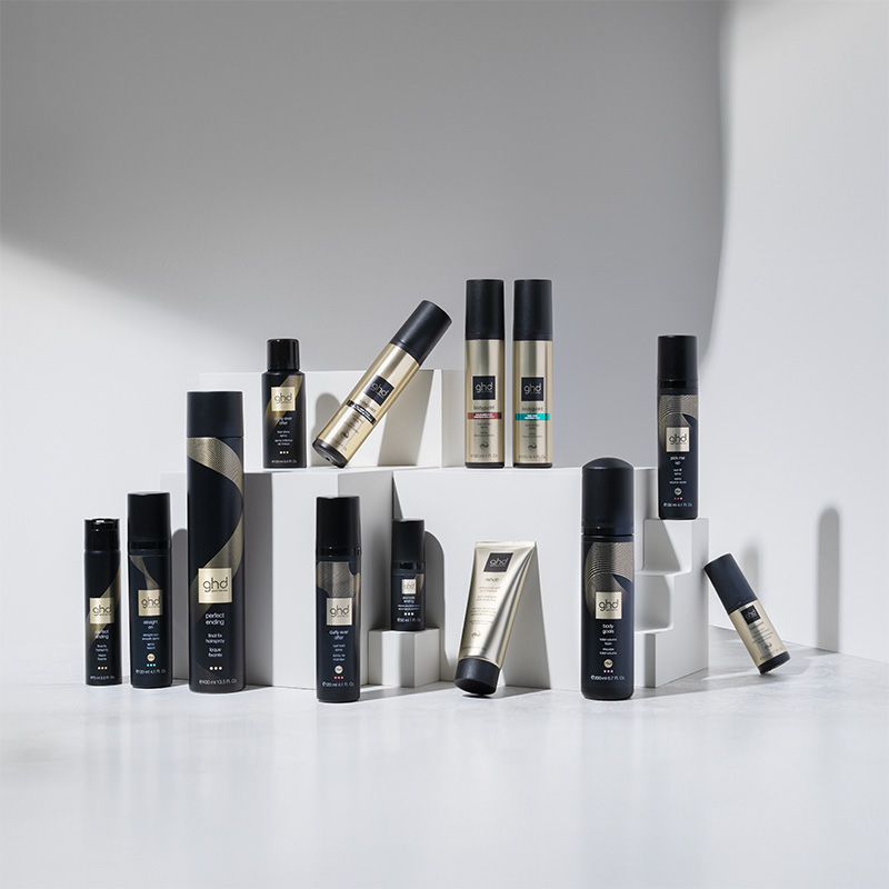 En samling av GHDs hårpleieprodukter er stilfullt arrangert på hvite kuber mot en lysegrå bakgrunn. Alle produktene kommer i elegante emballasjer i svart og gull. Lyset skaper skygger som fremhever produktenes design og plassering.
