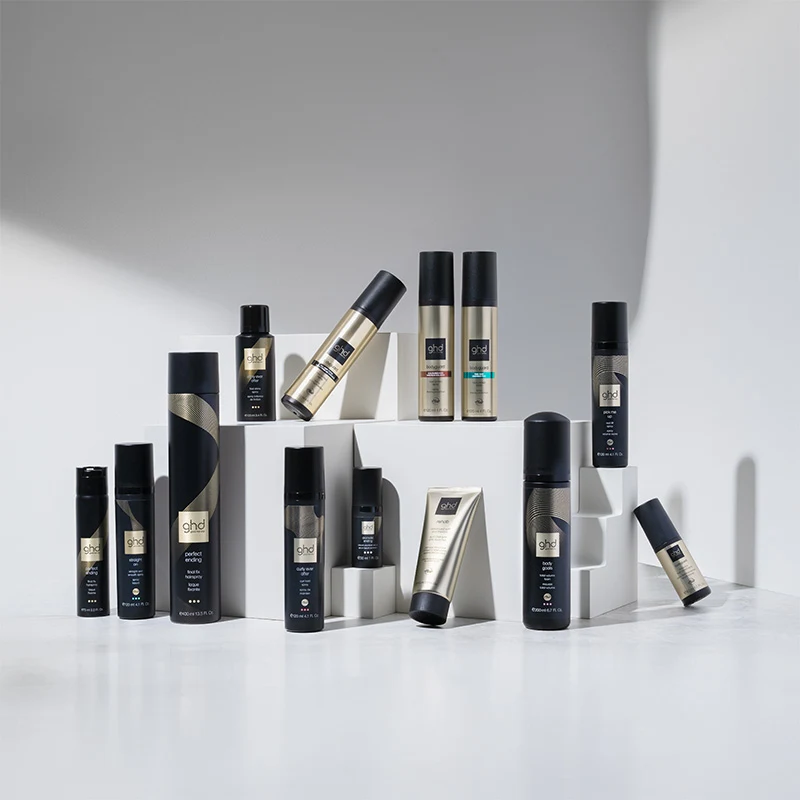 En samling GHD hårvårdsprodukter är stilfullt arrangerade på vita kuber mot en ljusgrå bakgrund. Produkterna är alla med eleganta svarta och guldiga förpackningar. Ljuset skapar skuggor som framhäver produkternas design och placering.