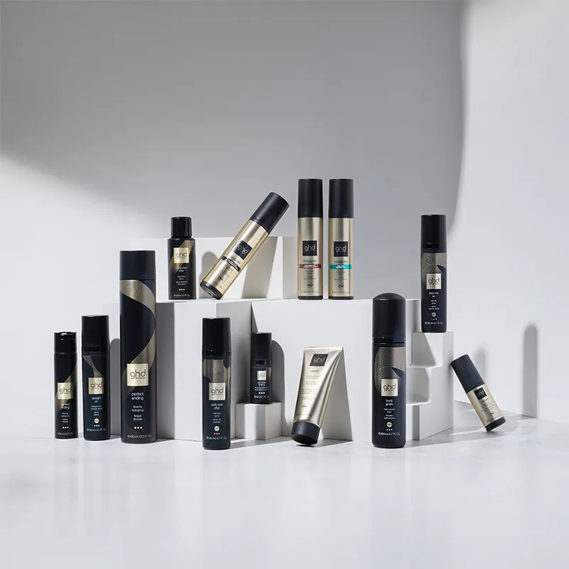 En samling GHD hårvårdsprodukter är stilfullt arrangerade på vita kuber mot en ljusgrå bakgrund. Produkterna är alla med eleganta svarta och guldiga förpackningar. Ljuset skapar skuggor som framhäver produkternas design och placering.