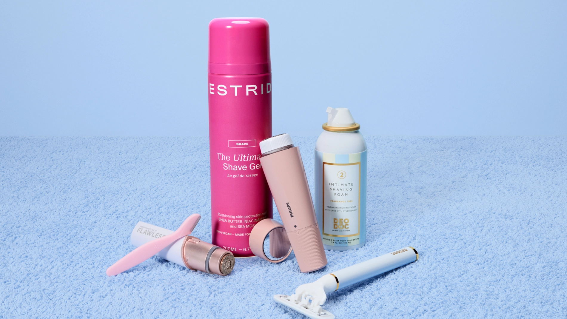 Sheivaus- ja intiimihygieniatuotteita esillä vaaleansinistä taustaa vasten, mukana muun muassa Estrid The Ultimate Shave Gel, DeoDoc Intimate Shaving Foam Fragrance Free, Philips sähkötrimmeri ja Flawless-karvanpoistolaite. Tuotteet on esitetty vaaleanpunaisissa, valkoisissa ja pastellisävyisissä pakkauksissa yhdessä höylän ja grooming-välineiden kanssa intiimialueen sheivaukseen ja ihonhoitoon.