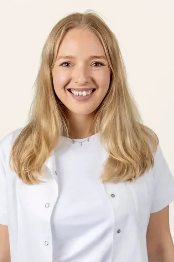 Portrett av en smilende person med langt blondt hår og hvit arbeidsjakke over hvit topp mot lys bakgrunn. Personen har naturlig makeup og et smykke rundt halsen.