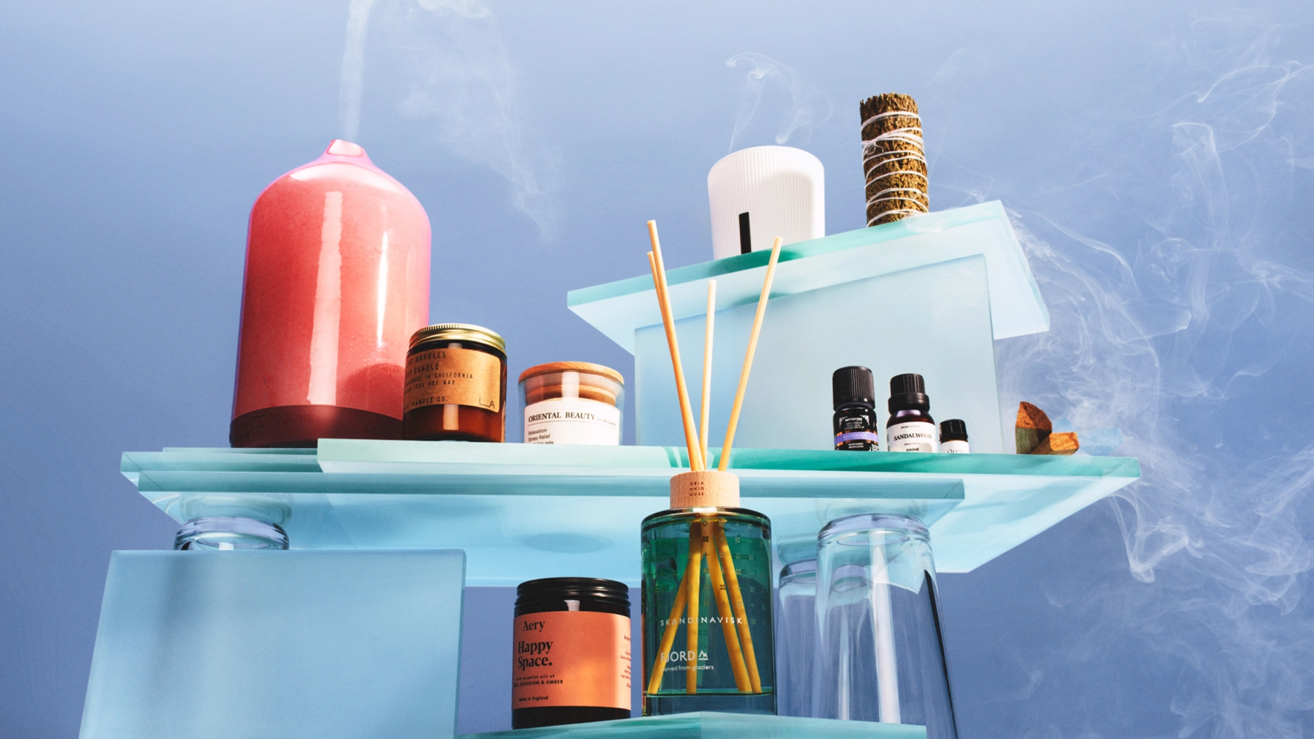 Hemdoft och wellnessprodukter uppställda på transparenta glasplattformar mot blå bakgrund med rökdetaljer. Bilden visar diffusern SKANDINAVISK FJORD med doftpinnar, doftljus från Aery Happy Space och Oriental Beauty, eteriska oljor inklusive “Sandalwood”, en rosa diffuser samt en bunt salvia.