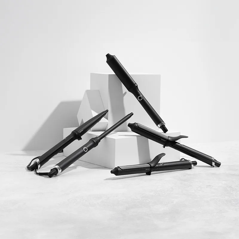 En samling svarta GHD-locktänger är stilfullt arrangerade på vita kuber mot en ljusgrå bakgrund. Locktängerna har eleganta detaljer och är placerade i olika vinklar för att visa deras design och funktioner.