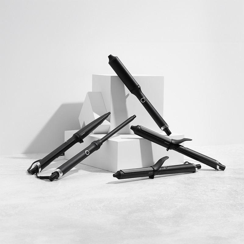 En samling svarte GHD-krølltang er stilfullt arrangert på hvite kuber mot en lysegrå bakgrunn. Krølltangene har elegante detaljer og er plassert i ulike vinkler for å vise design og funksjon.