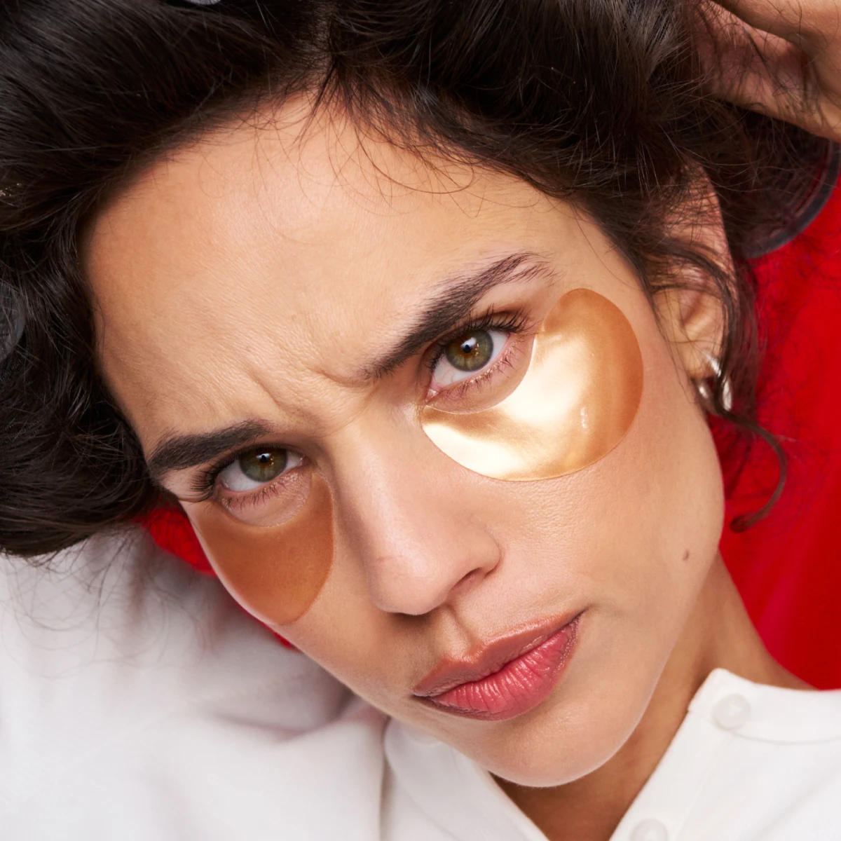 Kvinna med mörkt hår uppsatt med pappilliotter och guldiga eyepatches mot röd bakgrund. 
