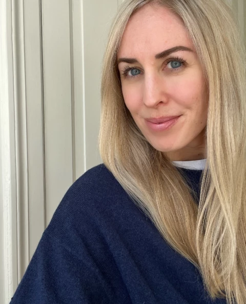 Person med langt blondt hår og mørkeblå genser. Bakgrunnen er en lys beige vegg.