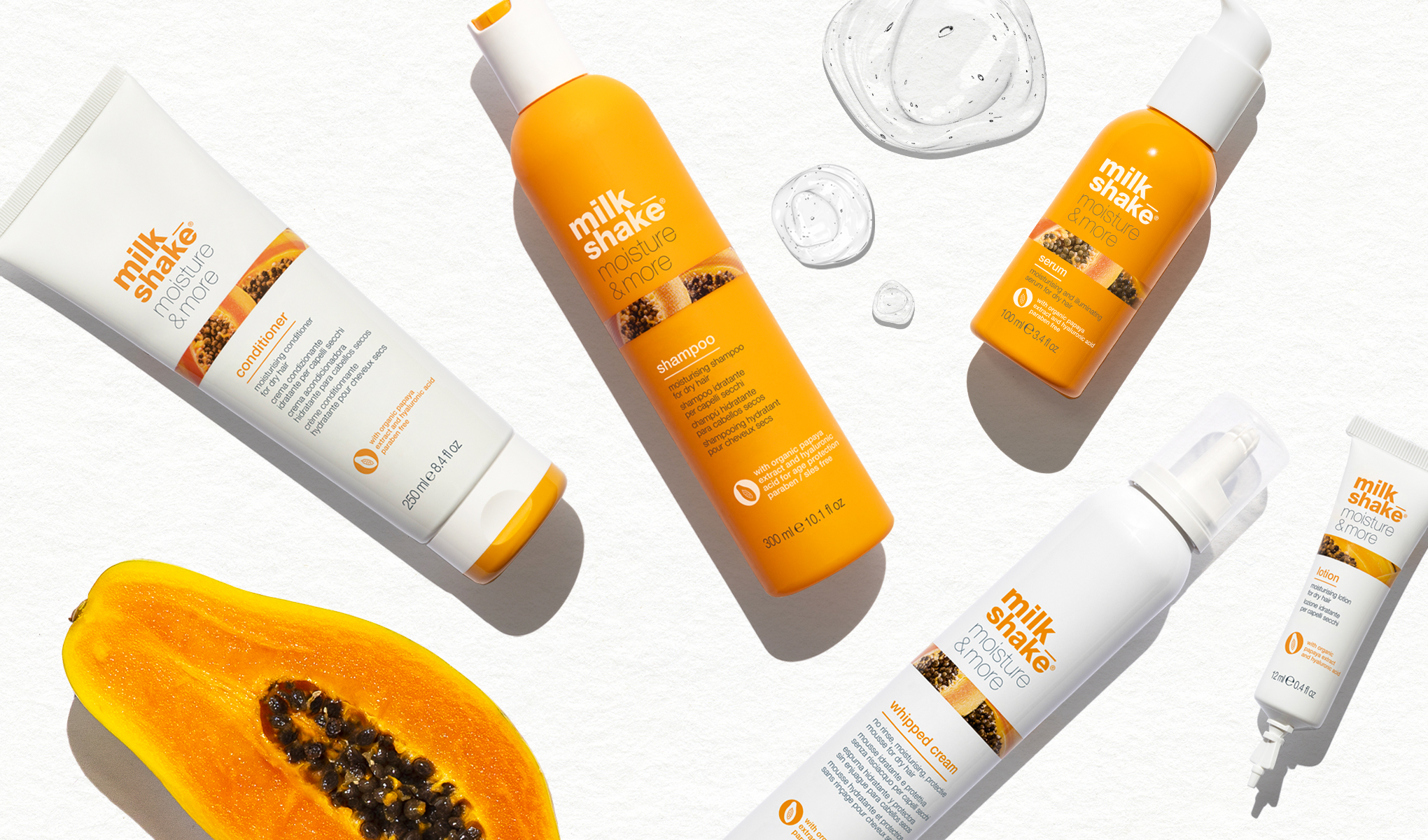 Flere Milk Shake-produkter, som sjampo, balsam, serum, skum og krem, plassert på en hvit bakgrunn. Bildet inkluderer også en halv papaya.