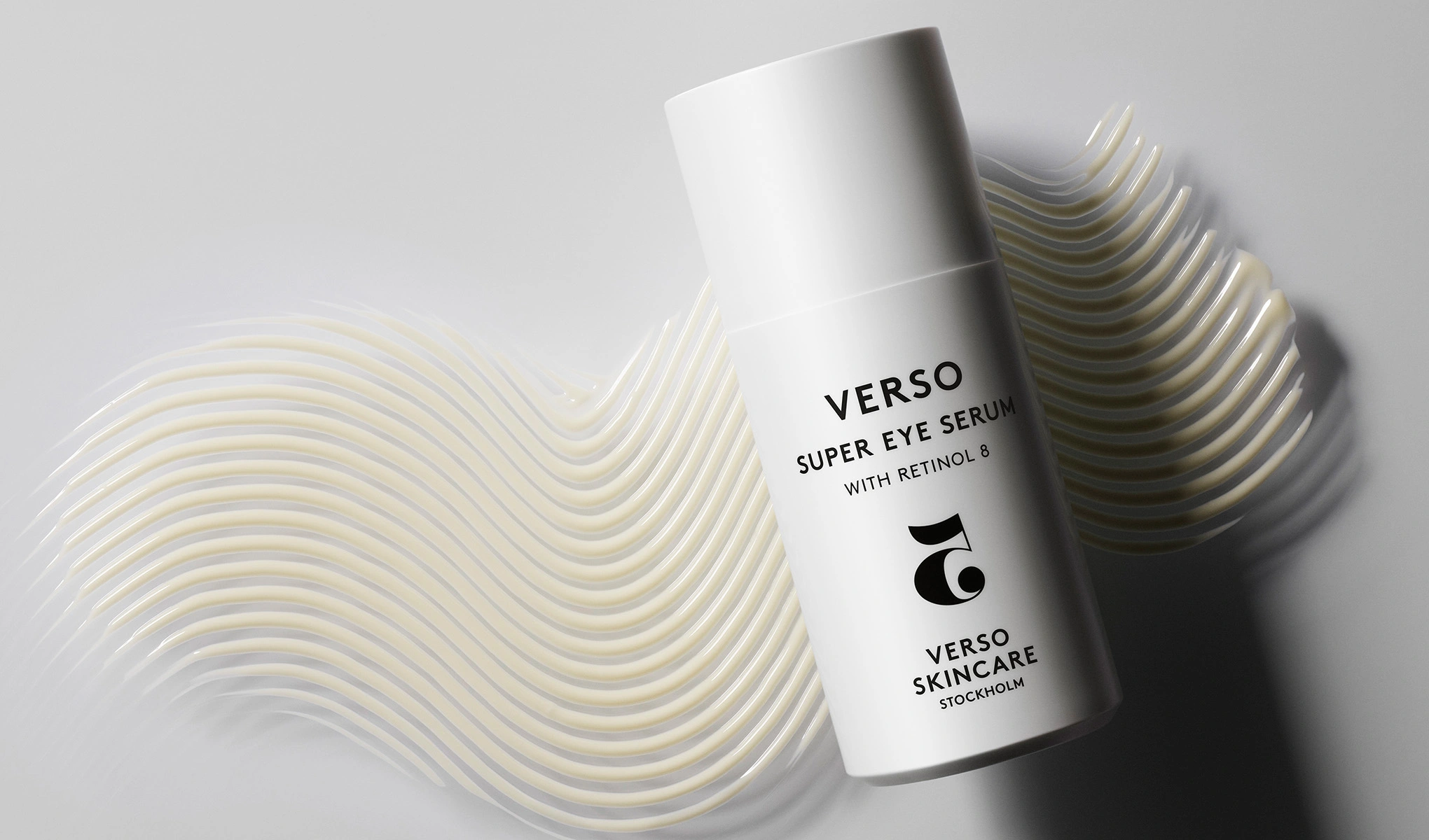 Verso Super Eye Serum, jossa on Retinol 8, valkoisessa pullossa. Pullo on asetettu vaaleanharmaalle taustalle, jossa on aaltoilevia, kermaisia viivoja. Viivat luovat tekstuurin taustalle ja korostavat tuotetta.