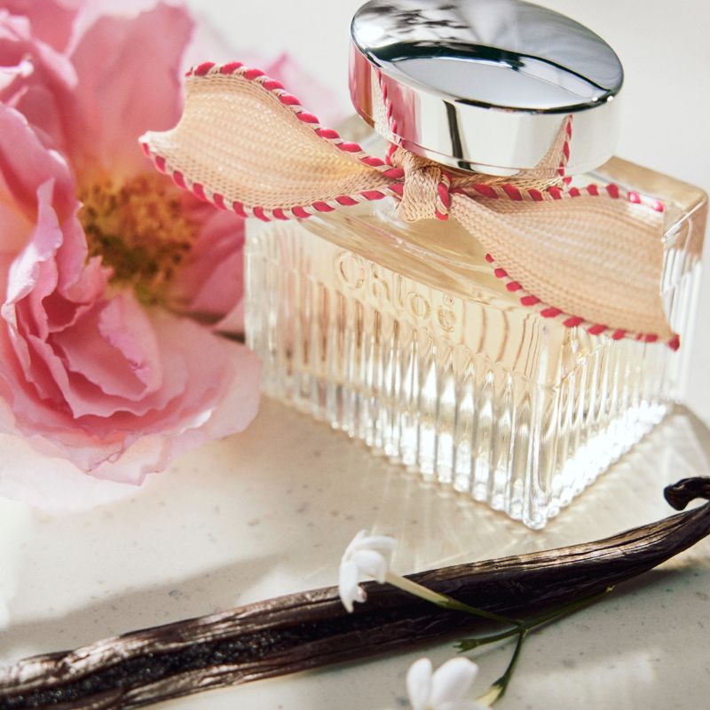 Parfymflaska från Chloé sin ör dekorerad med en beige rosett med röda kanter. Flaskan är placerad på en ljus yta tillsammans med en rosa blomma och en vaniljstång. Bakgrunden är ljus och mjukt belyst.