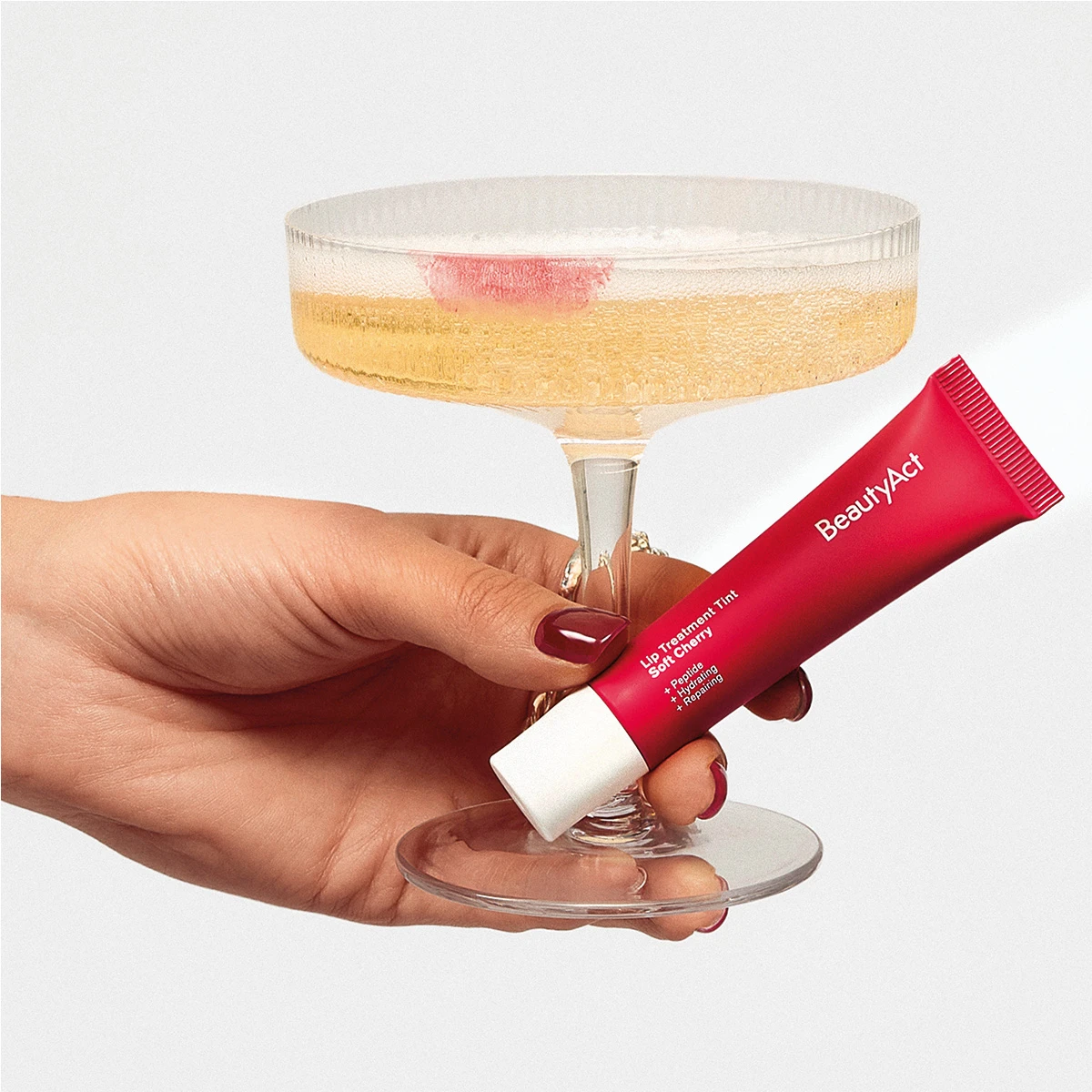 En hand håller en röd BeautyAct Lip Treatment Tint Soft Cherry-tub, med texten Peptide, Hydrating, Repairing. Bredvid står ett champagneglas med ett rött läppstiftsavtryck.