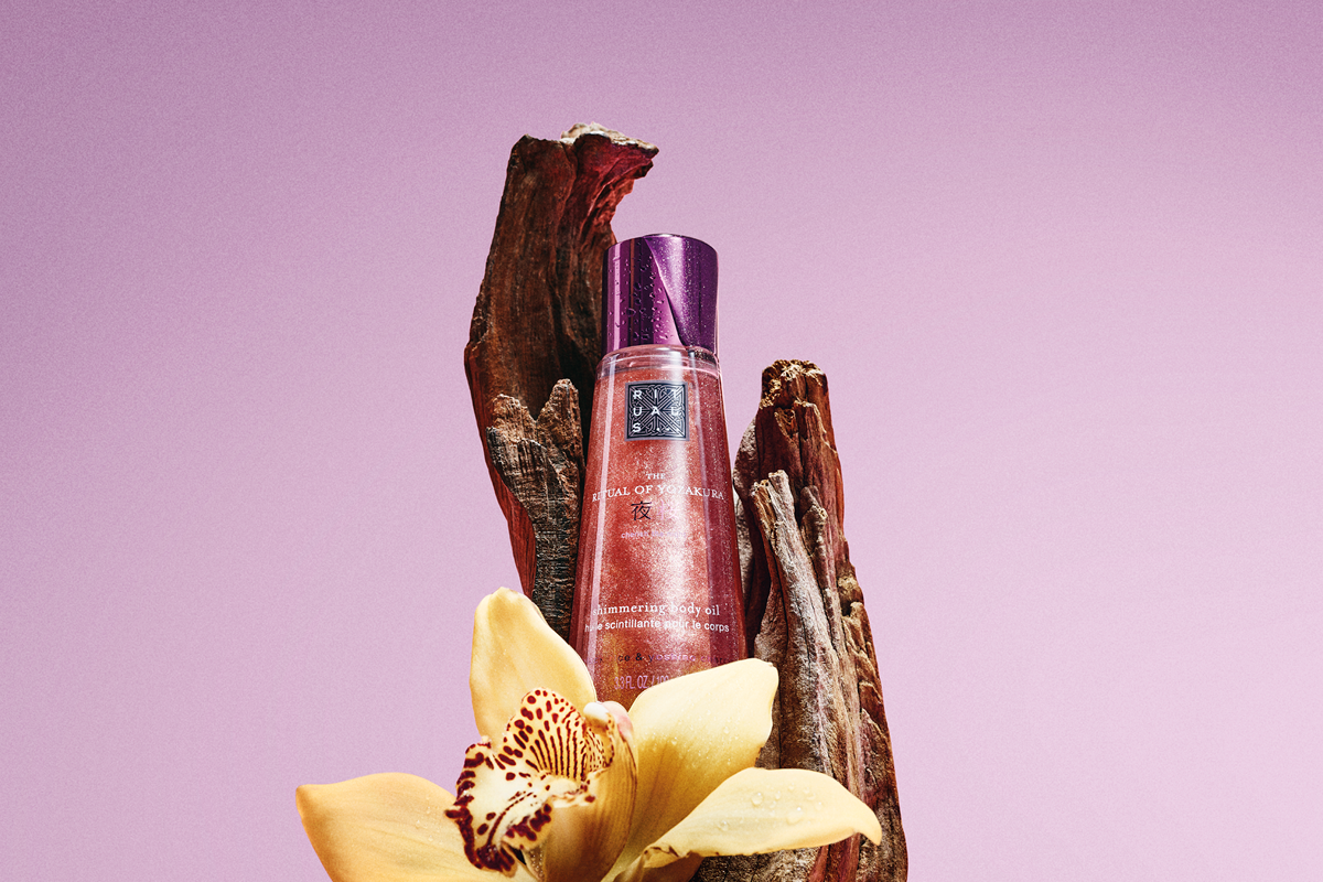 Flaska med Rituals The Ritual of Yozakura shimmering body oil står mellan träbitar och en gul blomma mot rosa bakgrund. Produktens etikett visar Rituals-logotypen, “THE RITUAL OF YOZAKURA” och “shimmering body oil”.