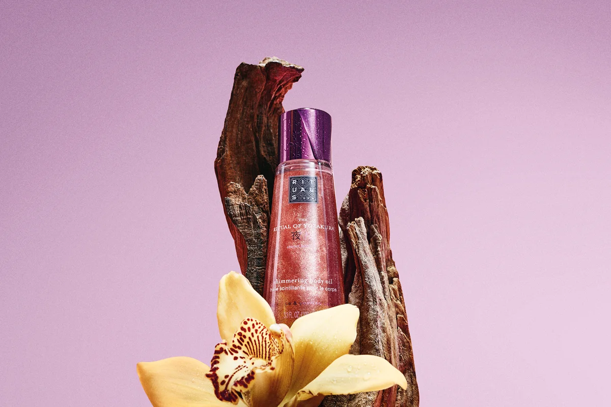 Flaska med Rituals The Ritual of Yozakura shimmering body oil står mellan träbitar och en gul blomma mot rosa bakgrund. Produktens etikett visar Rituals-logotypen, “THE RITUAL OF YOZAKURA” och “shimmering body oil”.