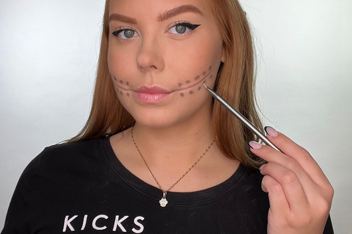 Kvinna med förlängd munmakeup, håller en sminkborste mot kinden. Prickar har lagts till med ögonskugga på båda sidor om munlinjen.