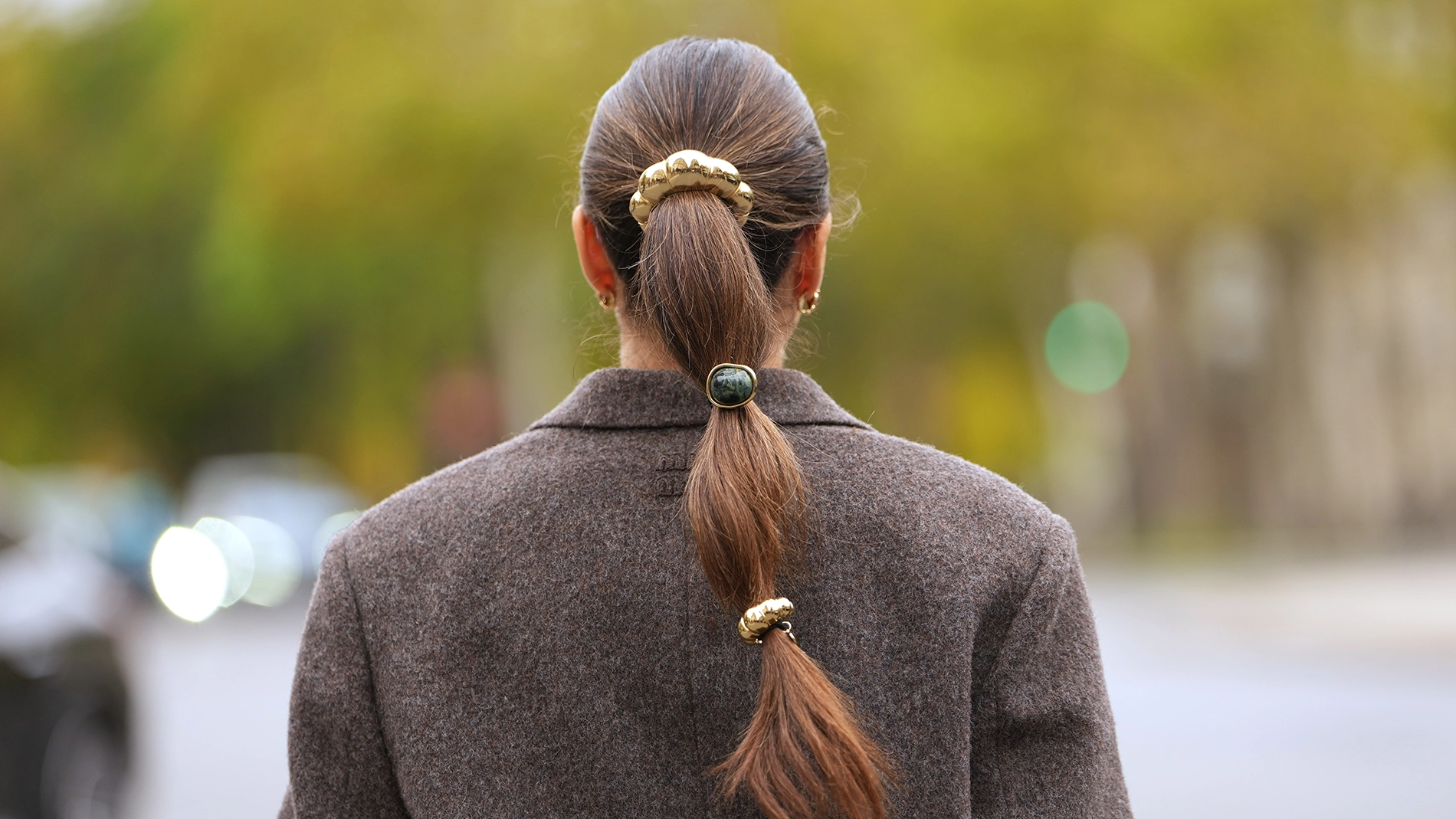 Henkilö nähtynä takaapäin, pitkät hiukset sidottuna matalaksi poninhännäksi useilla hiuslenkeillä. Henkilöllä on tumma takki ja hän seisoo ulkona kadulla, taustalla epätarkkoja puita ja liikennettä.