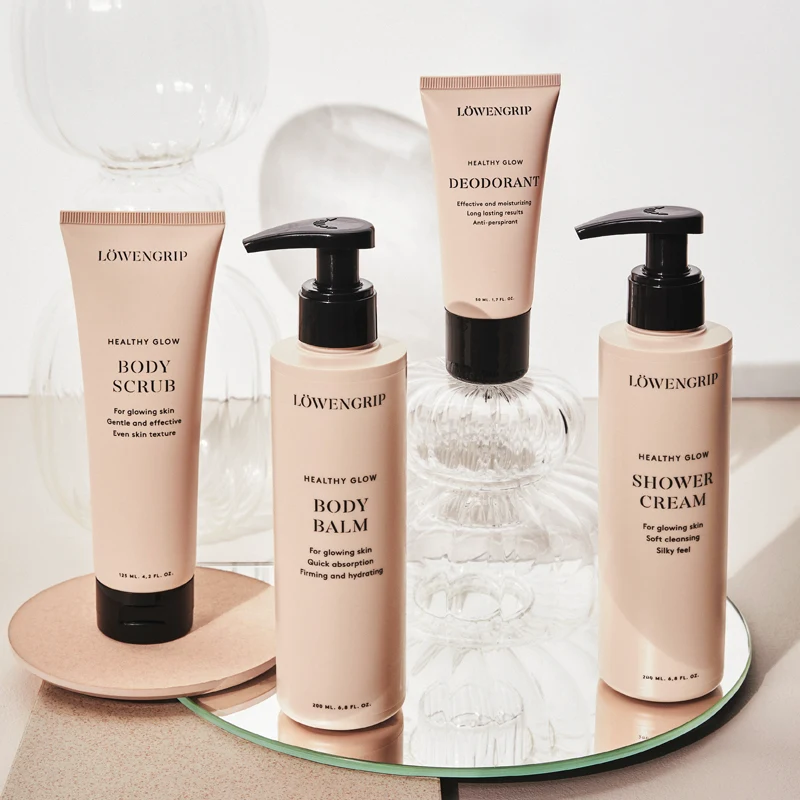 Fire kroppspleieprodukter fra Löwengrips Healthy Glow-serie er stilt opp mot en lys bakgrunn med dekorative glasskupler. Alle produktene har beige emballasje med svart tekst og svarte lokk. Produktene er plassert på et rundt speil og en beige plate.