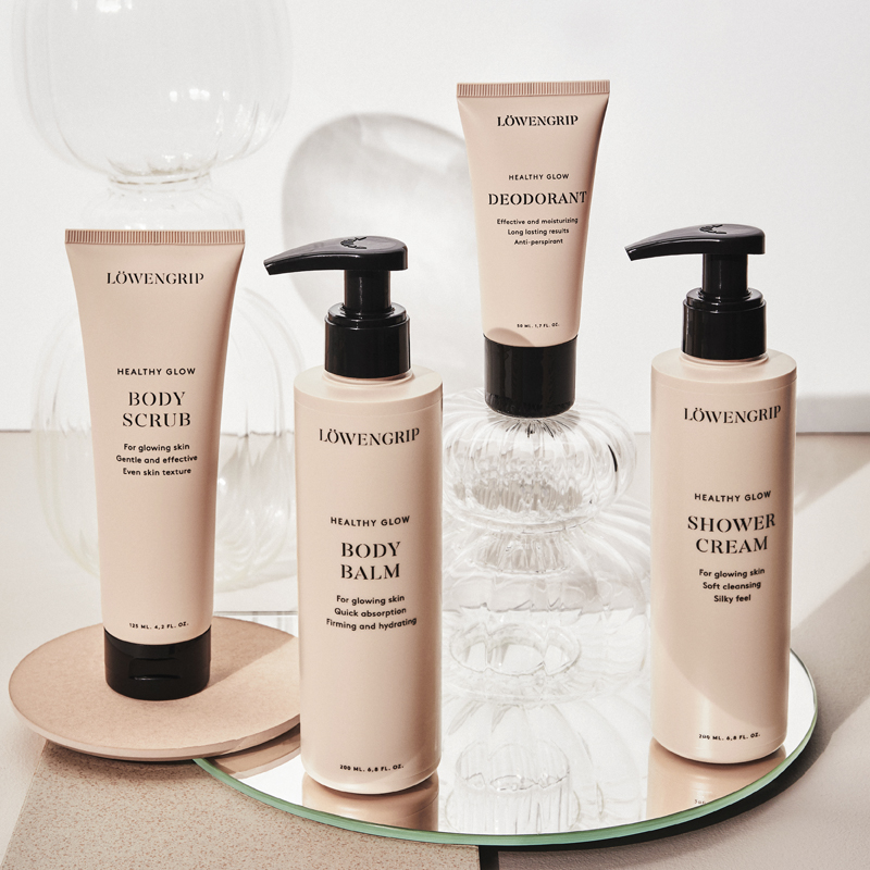 Neljä vartalonhoitotuotetta Löwengripin Healthy Glow -sarjasta on rivissä kirkkaalla taustalla, jossa on koristeellisia lasikupuja. Kaikissa tuotteissa on beige pakkaus, jossa on musta teksti ja mustat kannet. Tuotteet on sijoitettu pyöreän peilin ja beigen lautasen päälle.