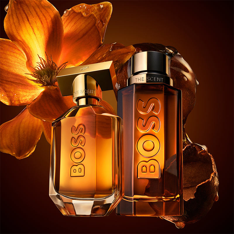 Hugo Boss The Scent-parfymflaskor i varma bärnstensfärger mot blomsterbakgrund.