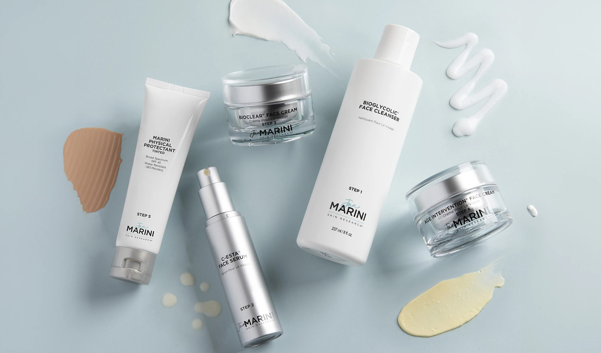 Flera hudvårdsprodukter från Jan Marini, placerade på en ljusblå bakgrund. Runt produkterna finns olika krämer och serum utsmetade på bakgrunden.