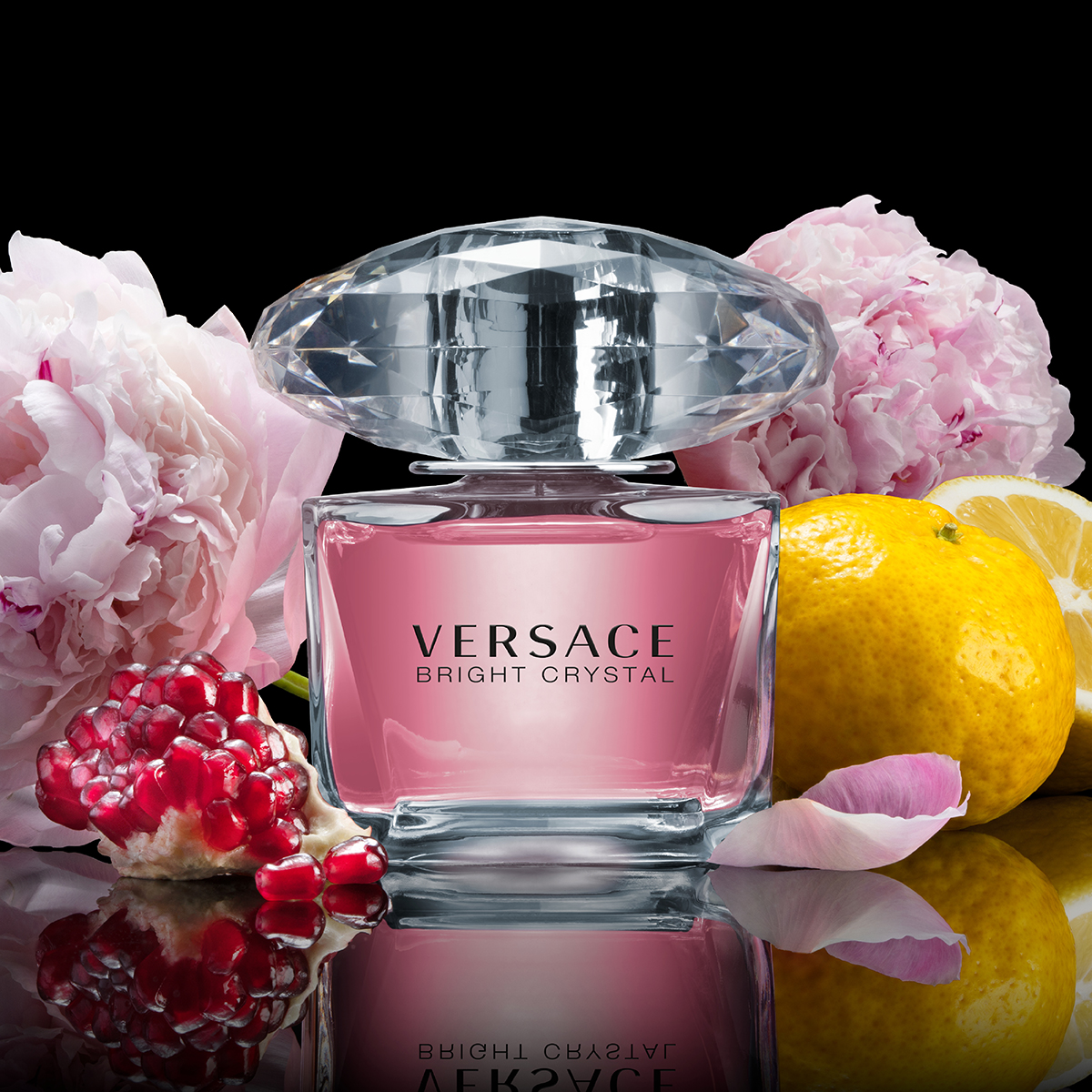 En rosa parfymflaska från Versace med en kristallklar kork, omgiven av rosa pioner, en citron, citronskivor och granatäpple mot en svart bakgrund. Texten "Versace Bright Crystal" syns på flaskan.