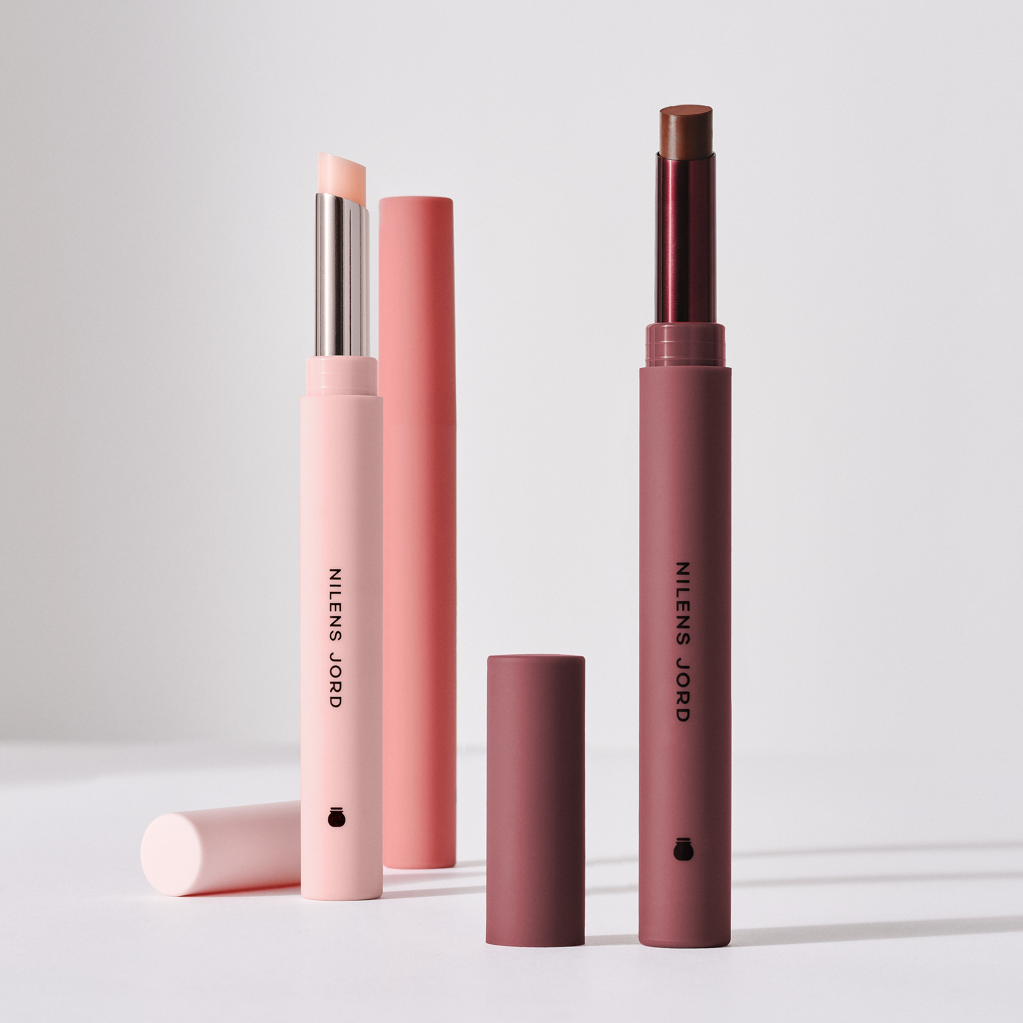 To åpne leppebalmer fra Nilens Jord i ulike nyanser med lokk ved siden av, arrangert mot en lys, nøytral bakgrunn.