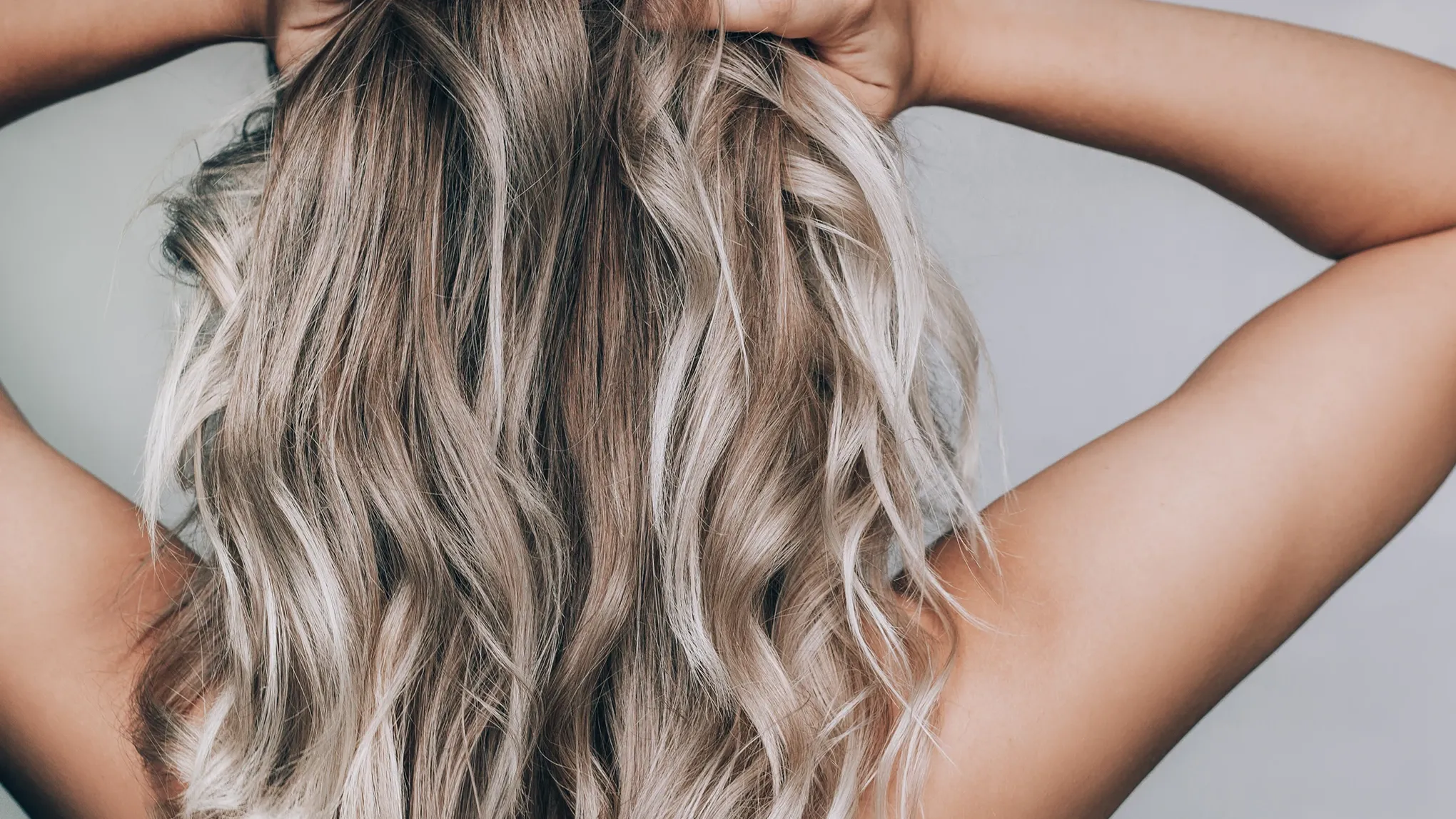  Närbild på en person med långt, vågigt blont hår som hålls upp med händerna. Håret har olika nyanser av blont och brunt, vilket skapar en naturlig och livfull look.