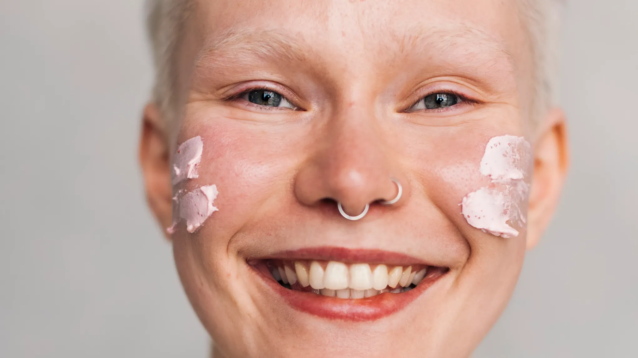 En person med kort, grått hår och två näspiercingar samt kräm på kinderna mot en grå bakgrund.