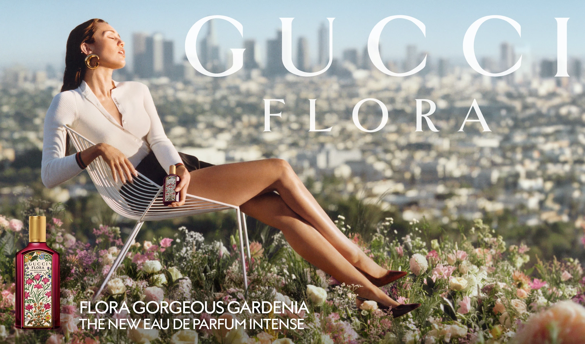 Nainen istuu tuolilla kukkien keskellä, pitäen kädessään Gucci Flora Gorgeous Gardenia -hajuvesipulloa. Taustalla näkyy kaupunkimaisema.