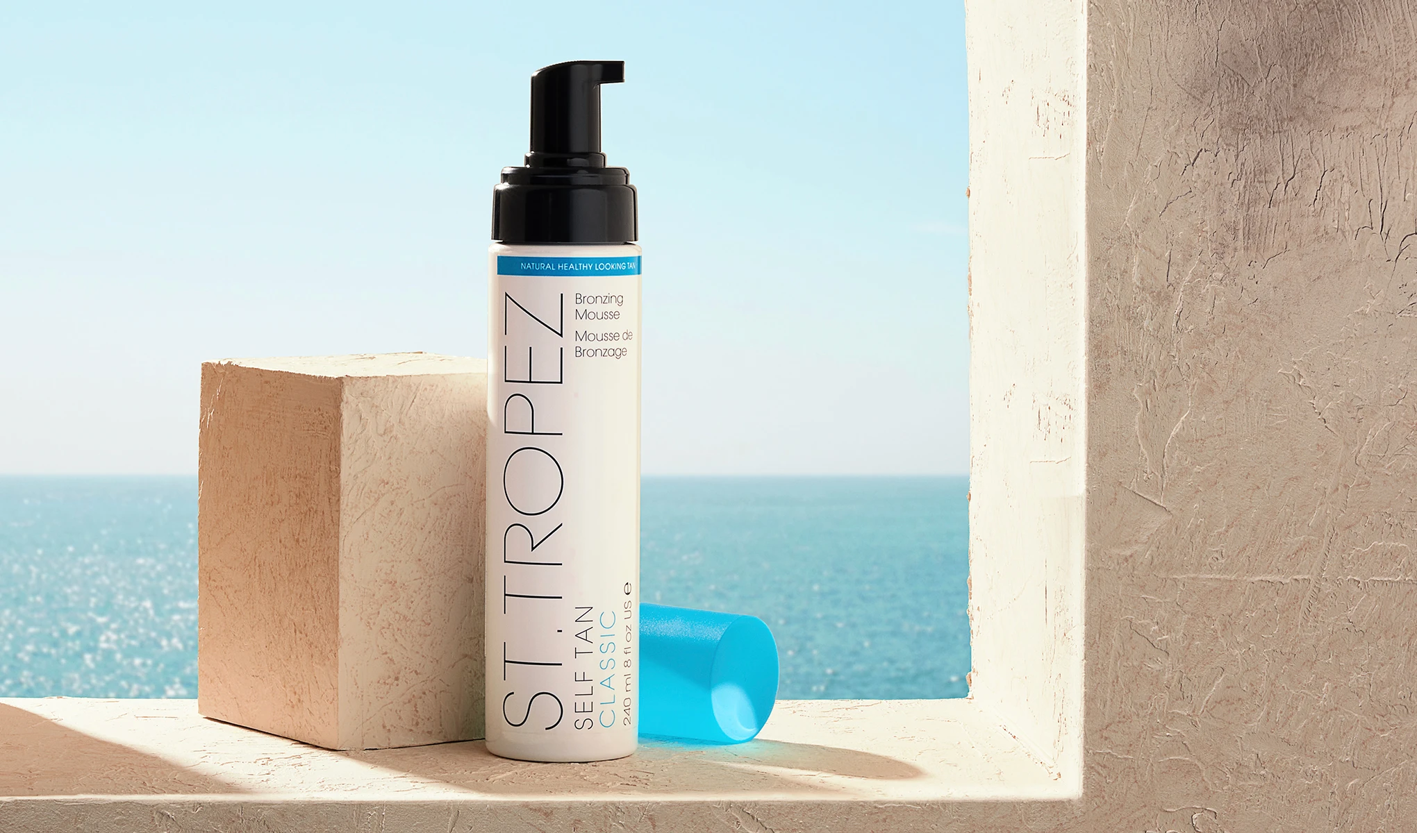 En hvit flaske med svart pumpe og blå kork fra St. Tropez plassert på en beige flate ved et vindu med utsikt over havet. Ved siden av flasken står en beige kube. Bakgrunnen viser en klarblå himmel og et glitrende hav.
