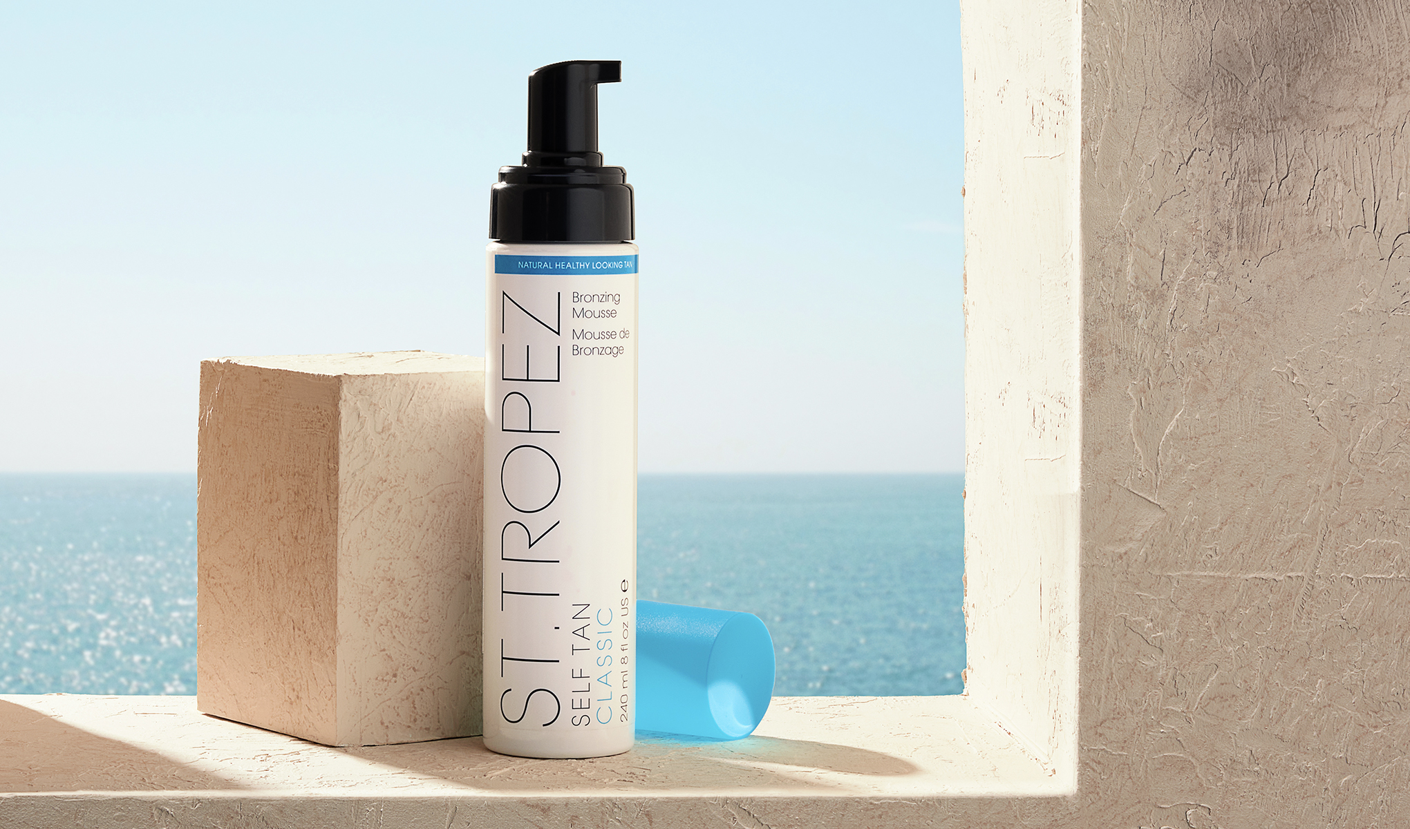 En vit flaska med svart pump och blå kork från St. Tropez placerad på en beige yta vid ett fönster med utsikt över havet. Bredvid flaskan står en beige kub. Bakgrunden visar en klar blå himmel och ett glittrande hav.
