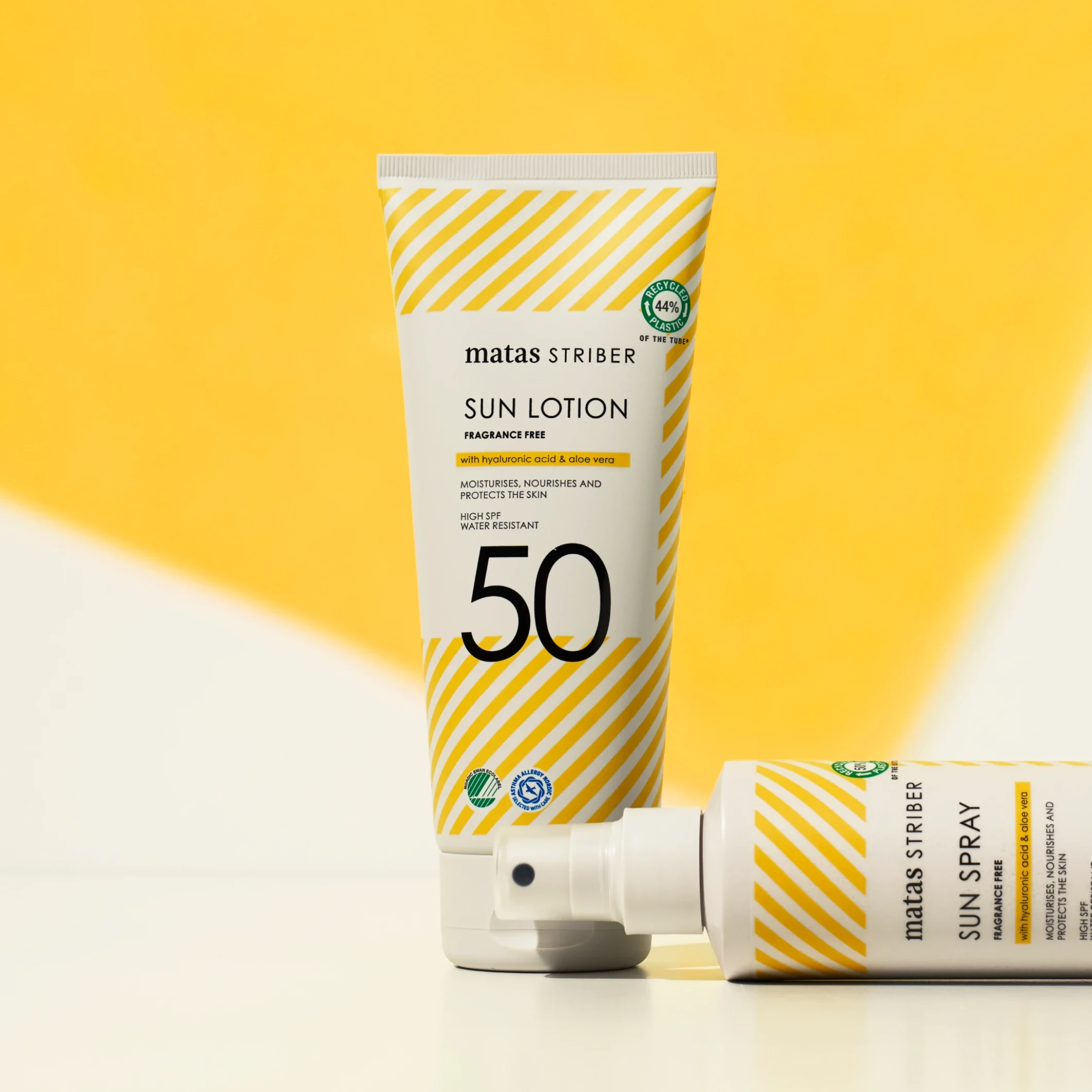 Matas Striber Sun Lotion SPF 50 står upprätt bredvid en Sun Spray-flaska som ligger horisontellt mot en gul bakgrund.