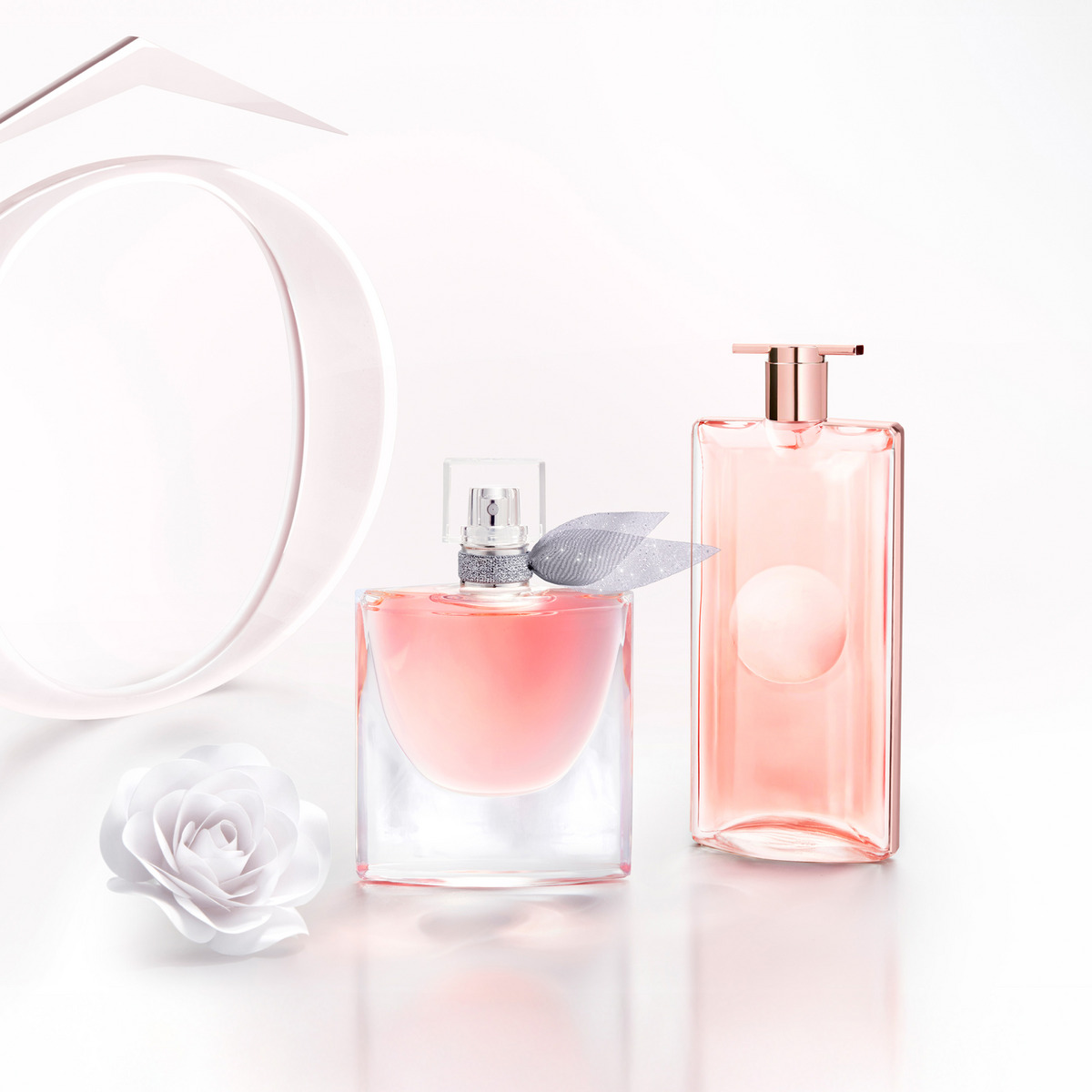 Kaksi vaaleanpunaista hajuvesipulloa vaalealla taustalla, toisessa hopeinen nauha kaulassa, vieressä valkoinen kukka.