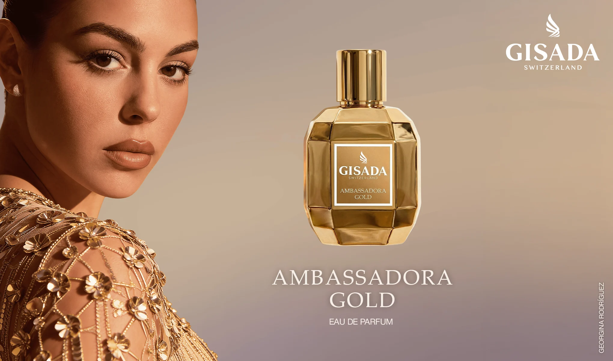 Gisada Switzerland Ambassadora Gold parfymflaska i guldfärgad design står på en skimrande gyllene stenplattform. I bakgrunden syns guldfärgade pelare, klippformationer, hav och berg i varmt ljus, samt texten "Gisada Switzerland" och etiketten "GISADA SWITZERLAND AMBASSADORA GOLD".