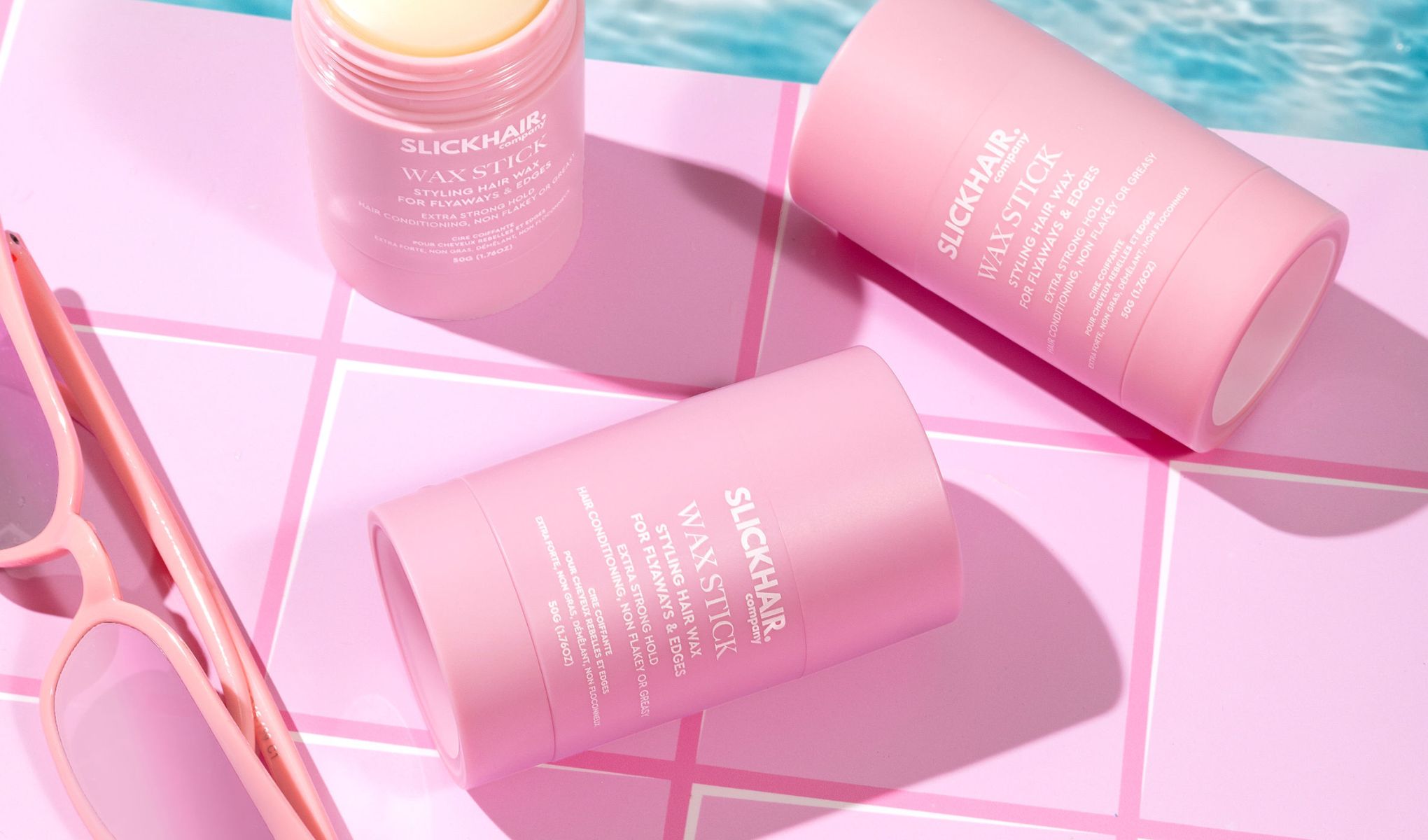 Tre rosa voksstifter fra merket Slickhair ligger på en rosa flislagt overflate ved siden av et par rosa solbriller. Bakgrunnen viser et svømmebasseng.