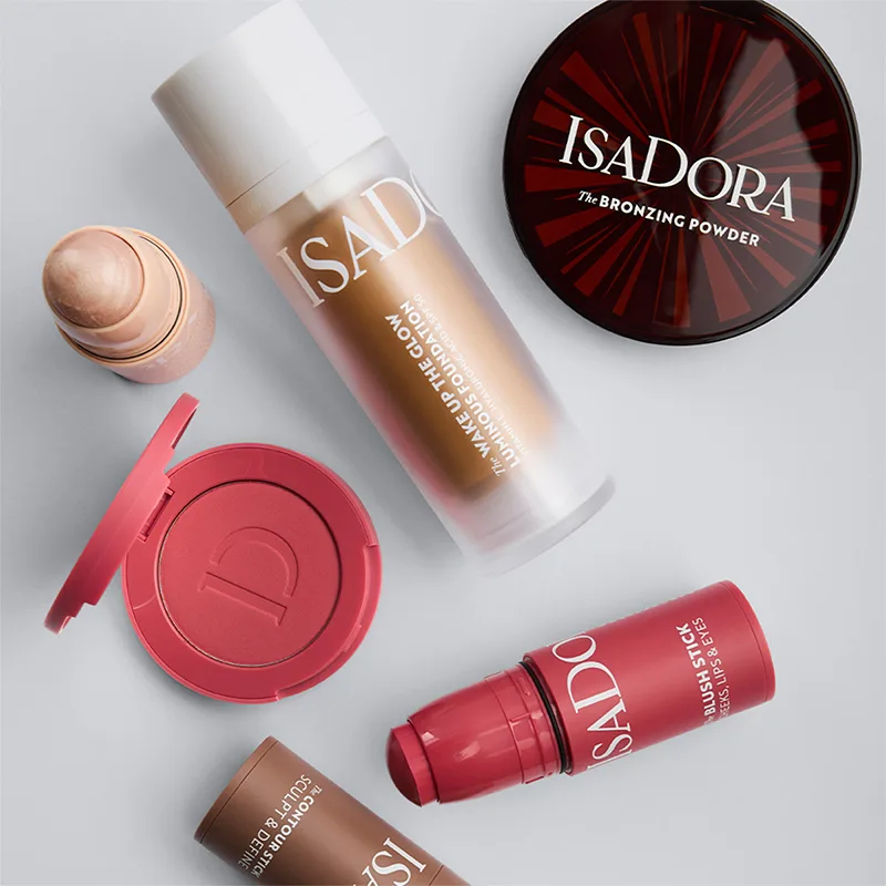 Flera IsaDora makeup-produkter inklusive The Bronzing Powder, The Wake Up the Glow Luminous Foundation och The Blush Stick arrangerade mot ljus bakgrund. Synliga texter på förpackningarna visar produktnamn och varumärke.