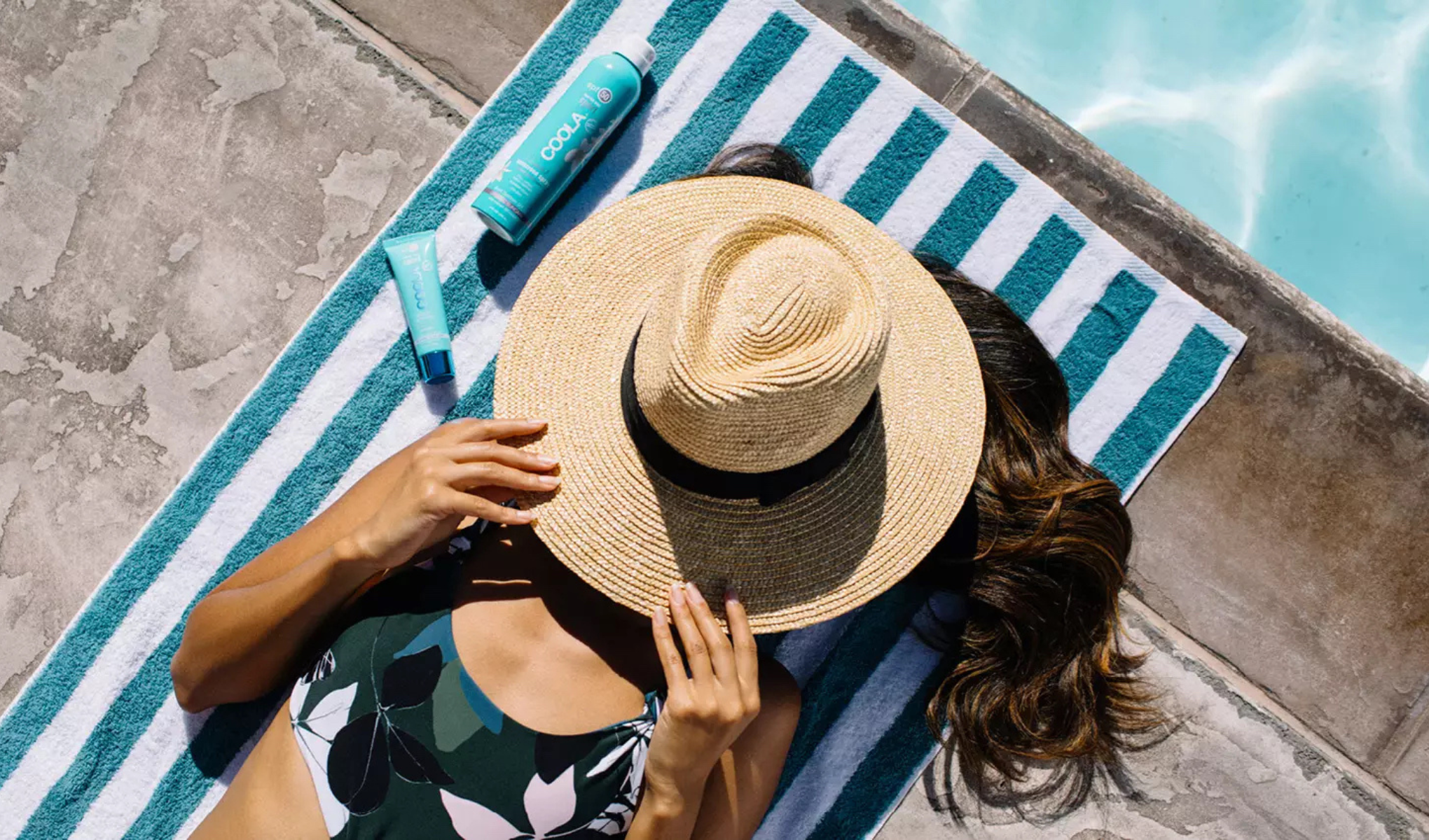 En kvinna ligger vid poolen på en randig handduk och håller en solhatt över ansiktet. Hon har på sig en blommig baddräkt. Bredvid henne ligger två solskyddsprodukter från märket Coola. Bakgrunden visar poolkanten och vattnet i poolen.