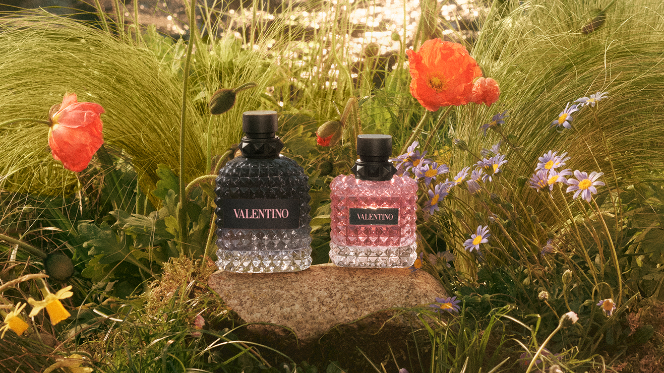 To Valentino-parfymeflasker står på en stein, omgitt av fargerike blomster og grønt gress.
