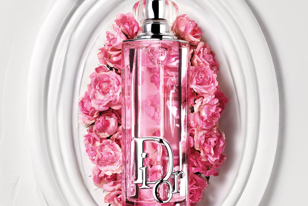 Dior Addict Lip Glow Oil i rosa flaska placerad på ett dekorativt vitt fat omgivet av rosa blommor.