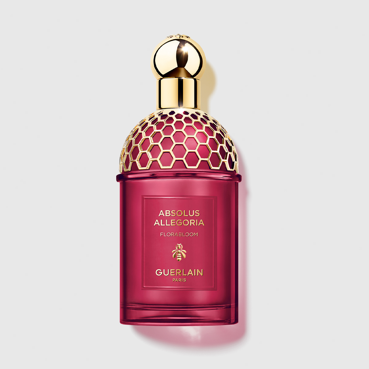 En röd parfymflaska med en guldfärgad kork med honungskakemönster står mot en ljus bakgrund. Flaskan har texten "ABSOLUS ALLEGORIA FLORABLOOM GUERLAIN PARIS".