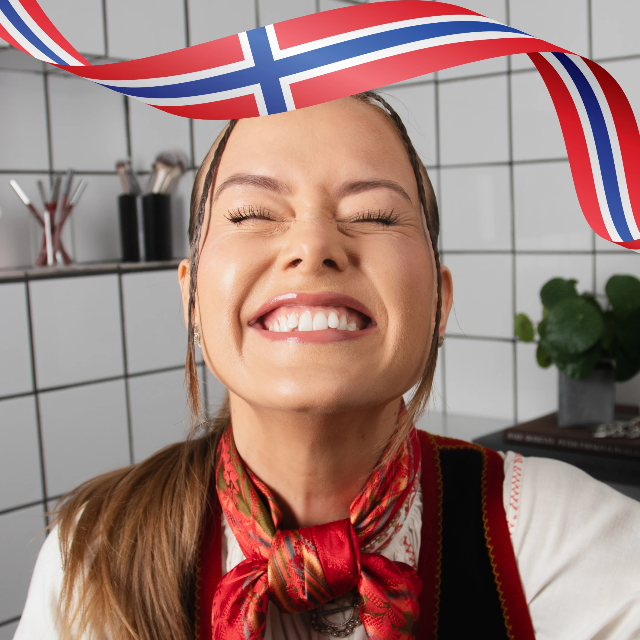 Person med oppsatt hår og rødt skjerf smiler med lukkede øyne i baderomsmiljø, med norsk flaggbånd over.