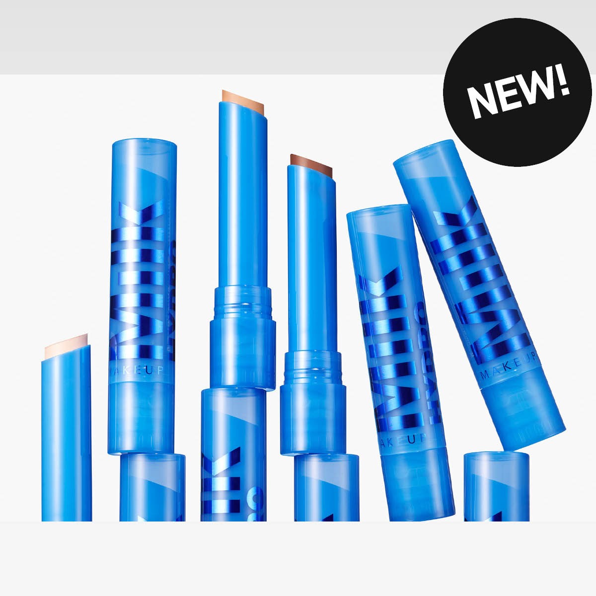 Useita sinisiä MILK MAKEUP -meikkipuikkoja avattuina eri kulmiin, näkyvissä beigejä ja ruskeita voidemaisia meikkisävyjä. Kulmassa näkyy musta ympyrä tekstillä “NEW!” ja pakkauksissa lukee “MILK MAKEUP”.