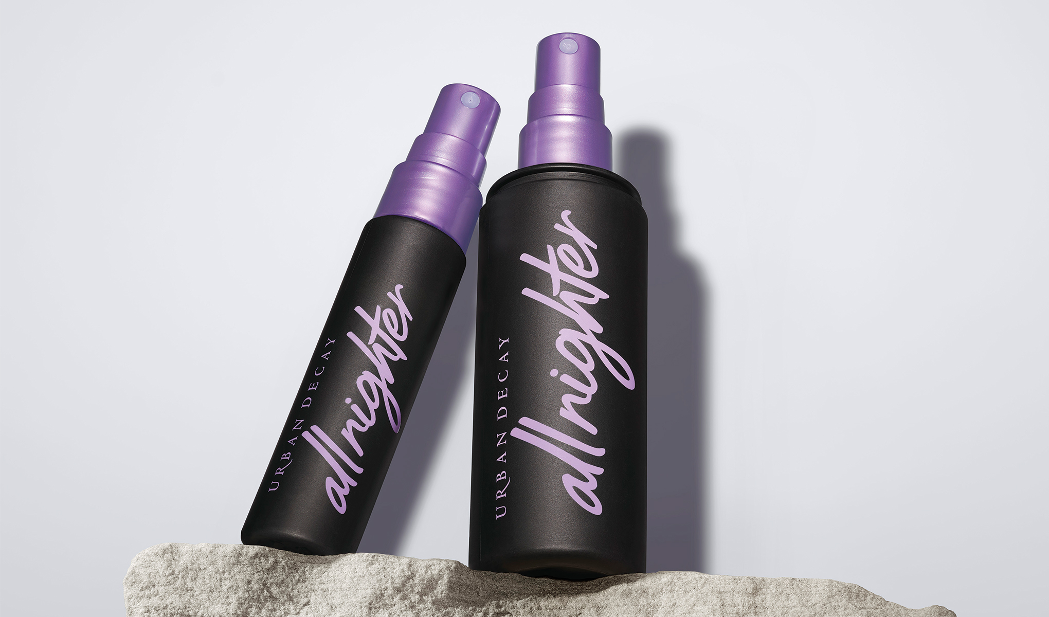 Kaksi Urban Decay All Nighter Setting Spray -pulloa, joissa on violetit suihkusuuttimet, seisoo kivisellä pinnalla valkoista taustaa vasten. Pullot ovat mustia, ja niissä on violetti teksti.