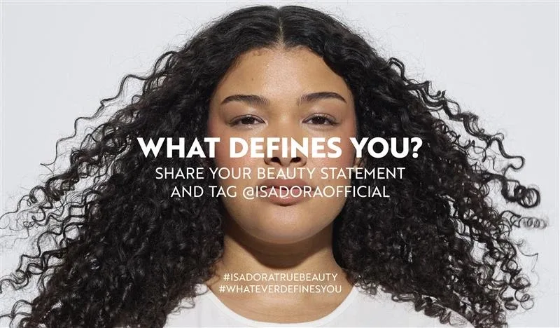 En person med lockigt hår står framför en vit bakgrund, med texten "WHAT DEFINES YOU? SHARE YOUR STATEMENT AND BECOME OUR SOCIAL" och hashtaggarna "#ISADORATRUEBEAUTY" och "#WHATEVERDEFINESYOU".