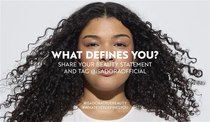 En person med krøllete hår står foran en hvit bakgrunn, med teksten "WHAT DEFINES YOU? SHARE YOUR STATEMENT AND BECOME OUR SOCIAL" og hashtagene "#ISADORATRUEBEAUTY" og "#WHATEVERDEFINESYOU".