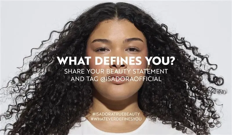 Henkilö, jolla on kiharat hiukset, seisoo valkoista taustaa vasten, tekstillä "WHAT DEFINES YOU? SHARE YOUR STATEMENT AND BECOME OUR SOCIAL" ja hashtageilla "#ISADORATRUEBEAUTY" ja "#WHATEVERDEFINESYOU".