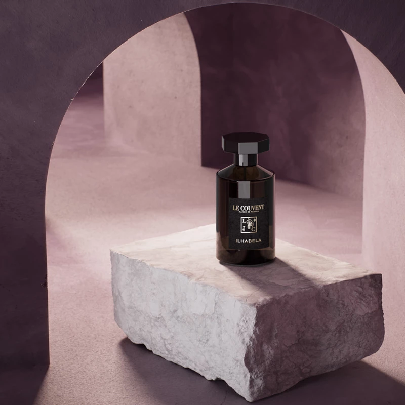 En mörk glasflaska med parfym märkt "Le Couvent Ilhabela" står på en ljus stenblock i ett lila rum med välvda väggar.